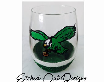 May include: A clear stemless wine glass featuring a green and white eagle graphic holding a football. The base of the glass is decorated with a band of white glitter above a wider band of green glitter. The text Etched Out Designs is below.