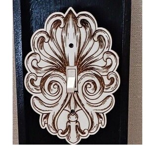 May include: A white light switch cover with a decorative, laser-cut design of intricate swirls and flourishes. The design is reminiscent of a stylized floral pattern.