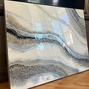 May include: Abstract art piece with a glossy finish. The artwork features flowing, layered designs in shades of white, gray, black, and gold. The piece has a textured appearance with embedded crystals and a signature in the bottom right corner.