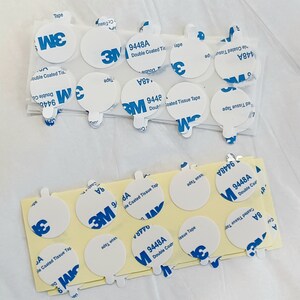 May include: A close-up of a stack of white, round, double-coated tissue tape dispensers with blue 3M branding and the product number 9448A. The tape dispensers are on a white surface.