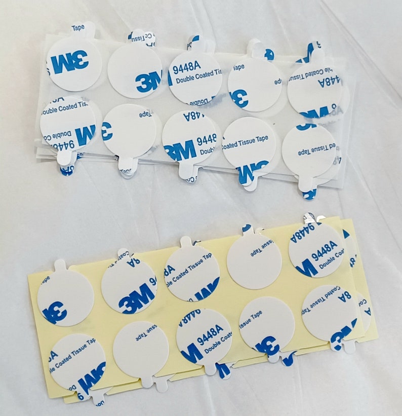 May include: A close-up of a stack of white, round, double-coated tissue tape dispensers with blue 3M branding and the product number 9448A. The tape dispensers are on a white surface.