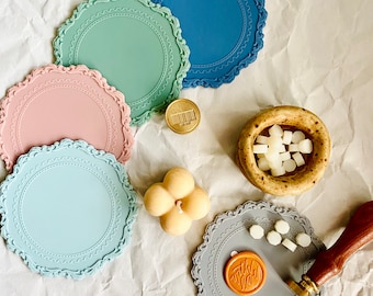 May include: Five colorful silicone coasters with a decorative scalloped edge. The coasters are in shades of blue, green, pink, and gray. A small, round, gold-colored metal object is in the center of the image. A small, white, four-leaf clover-shaped candle is in the lower right corner of the image. A small bowl filled with white wax pellets is in the upper right corner of the image. A gray silicone coaster with a decorative scalloped edge has a brown wax seal with the word "With Love" on it. A wooden handle for a wax seal is in the upper right corner of the image.