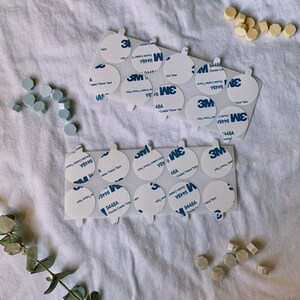 May include: A close-up of a sheet of white fabric with multiple round, white adhesive stickers. The stickers have the text "3M 9448A Double Coated Tissue Tape" printed on them. There are also several small, light blue and light beige ceramic tiles scattered around the stickers.