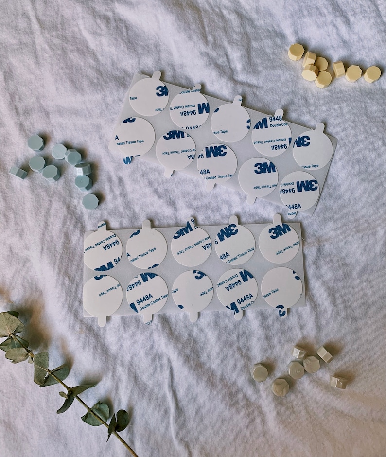 May include: A close-up of a sheet of white fabric with multiple round, white adhesive stickers. The stickers have the text "3M 9448A Double Coated Tissue Tape" printed on them. There are also several small, light blue and light beige ceramic tiles scattered around the stickers.