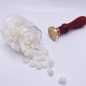 May include: A clear plastic jar filled with white wax sealing pellets, spilling out onto a white surface. A wooden handled wax seal stamp with a gold metal base is also pictured.