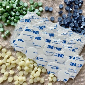 May include: An assortment of wax seal beads in shades of green, blue, and cream, alongside sheets of double-sided adhesive tape. The wax beads are hexagonal and cylindrical, perfect for crafting and stationery projects.