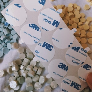 May include: A sheet of white adhesive tape with multiple round labels. The labels are white with blue text and a 3M logo. The text on the labels reads "Double Coated Tissue Tape" and includes a product number. The labels are arranged in rows and columns. There are also a variety of small, hexagonal wax seals in white and light blue.