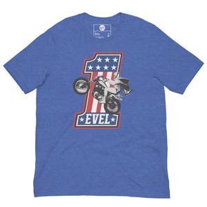 May include: Blue t-shirt with a graphic of a motorcycle stunt rider and the number one in red and white, with the word "EVEL" in a star-bordered box. The design has a vintage, patriotic aesthetic.