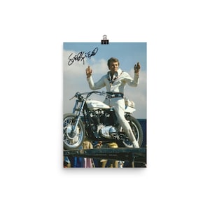 May include: A black and white photo of a man in a white jumpsuit performing a stunt on a Harley Davidson motorcycle. The man is raising his arms in the air and the motorcycle is balanced on a ramp. The photo is signed "Evel Knievel".