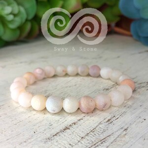 May include: A white agate beaded bracelet with a frosted finish. The beads are round and have a slight pink hue. The bracelet is on a white wooden surface.