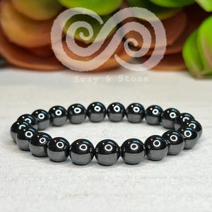 May include: A dark gray beaded bracelet with a glossy finish. The bracelet is made of round, polished beads strung together. The background features a blurred arrangement of green and orange foliage, with the text "Sway & Stone" in the upper center.