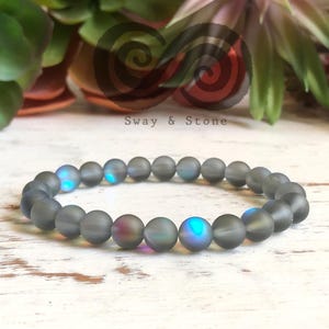 May include: A gray beaded bracelet with iridescent beads. The beads have a frosted finish and shimmer with blue and green hues.