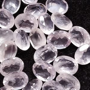 Puede incluir: Una colección de gemas ovaladas de color rosa pálido. Las gemas están facetadas, captando la luz y mostrando un sutil brillo. Las piedras están dispersas sobre una superficie oscura, creando un contraste que resalta su color y claridad.