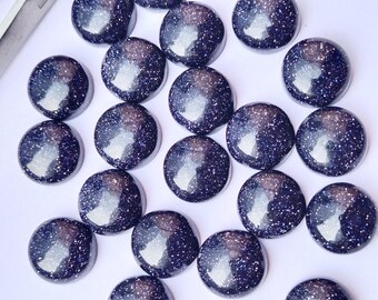 May include: A collection of round, polished blue sandstone cabochons with sparkling inclusions. These gemstones are arranged on a white surface, showcasing their deep blue color and reflective qualities. Ideal for jewelry making.