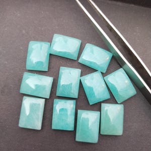 May include: A collection of ten rectangular, light blue gemstones with polished surfaces. The stones are arranged on a dark surface, with a pair of silver tweezers in the background. The gemstones have a subtle, natural pattern.