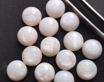 May include: A collection of luminous, white, round cabochon gemstones. The gemstones have a pearlescent sheen and are arranged on a dark surface. A pair of silver tweezers is positioned near the gemstones.
