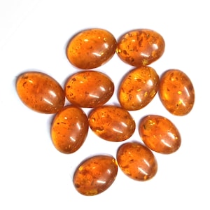 May include: A selection of oval amber gemstones with a polished finish. The stones, in varying sizes, display warm orange and brown tones with inclusions. They are arranged on a white background, showcasing their natural beauty.