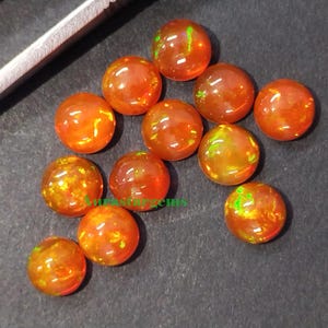 May include: A selection of round, orange-red opal gemstones with internal colour flashes. The opals are scattered on a dark surface, with tweezers visible. The gemstones have a polished, glossy appearance. Each gem is approximately 0.8 cm in diameter.