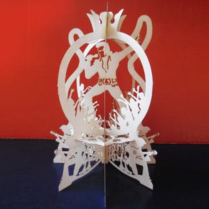 May include: A white paper sculpture depicting a performer with a crown, encircled by a crowd. The sculpture is within a circular frame with decorative details. The performer holds a microphone, surrounded by fans. The base of the sculpture reads "Rock and Roll".