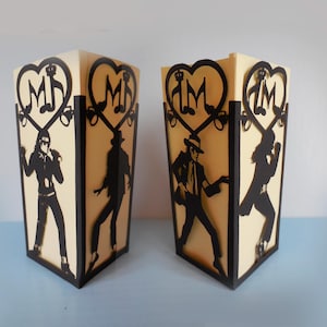 Puede incluir: Dos cajas decorativas con siluetas negras de un cantante en varias poses. Las cajas son de color crema con ribetes negros y presentan un diseño en forma de corazón con notas musicales y las iniciales "MJ".