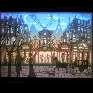 May include: A paper cut-out art piece depicting a Parisian street scene with the Moulin Rouge cabaret in the center. The scene is lit from behind, creating a glowing effect. Silhouettes of people and a horse-drawn carriage are visible in the foreground.