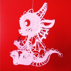 May include: A pink papercraft dragon with intricate details, including wings, a tail, and a large eye. The dragon is suspended in mid-air, as if it is flying.