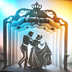 Puede incluir: Una silueta de papel blanco de Cenicienta y el Príncipe Azul bailando bajo un arco de papel con la palabra "Cenicienta" escrita en él. El arco está decorado con estrellas, corazones y un reloj.