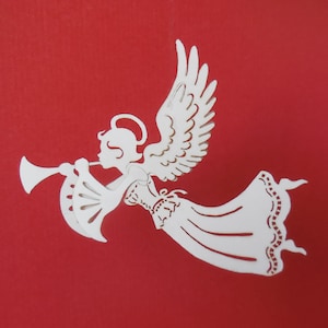 May include: White paper cut-out of an angel playing a trumpet. The angel has a halo and flowing robes. The background is red.