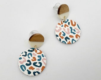 May include: A pair of dangle earrings with a white and brown wooden top and a white circle with a pink, blue, and orange abstract pattern.