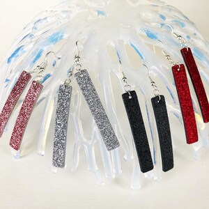 May include: Six pairs of dangle earrings with silver hooks and rectangular glitter-covered charms. The charms are in red, black, and silver.