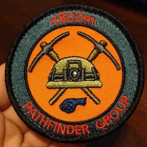 May include: A round, orange and blue embroidered patch with the text "Pathfinder Group" and "1800m" in red. The patch features a crossed pickaxe and a miner's helmet.