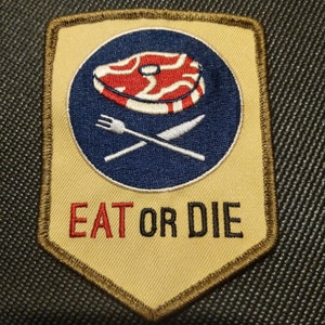 May include: A tan and brown embroidered patch with a blue circle containing a red and white steak, a fork and knife, and the text "EAT OR DIE".