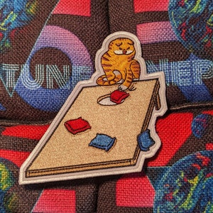 May include: A brown and white cat with orange eyes is playing a game of cornhole. The cat is standing on the cornhole board and is about to throw a red bean bag. The board is made of wood and has a white border. There are two red bean bags on the board and one blue bean bag.