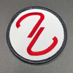 May include: A white circular patch with a red embroidered letter "Z" in the center. The patch has a black border.