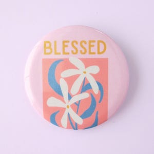 May include: A round, pink button with the word "BLESSED" in gold letters at the top. The center features a pink square with white flowers, blue curved lines, and a light pink background.