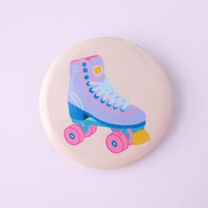 May include: A round, cream-colored button featuring a colorful illustration of a retro roller skate. The skate is light purple with blue, pink, and yellow accents. The button is a fun, nostalgic accessory.