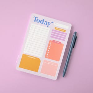 May include: A lavender daily planner with the word "Today" in blue script. The planner includes sections for priorities, to-do lists, and notes, with orange and pink accents. A blue pen rests beside the planner on a matching lavender surface.
