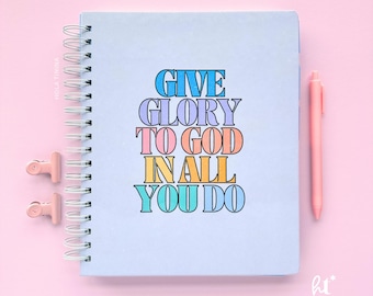Puede incluir: Un cuaderno azul claro con espiral con el texto "GIVE GLORY TO GOD IN ALL YOU DO" en letras de bloque coloridas. Un bolígrafo rosa y dos clips rosas también son visibles sobre un fondo rosa.