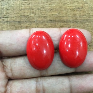 Könnte beinhalten: Zwei leuchtend rote, ovale Cabochons, wahrscheinlich Edelsteine, werden auf einer Hand gezeigt.
