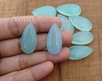 Op de afbeelding: Een close-up van tien traanvormige, lichtblauwe chalcedoon edelstenen. De edelstenen zijn glad en gepolijst en liggen in een stapel op een houten oppervlak.