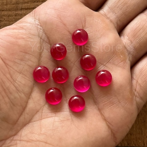 May include: Ten round, bright pink cabochon gemstones, likely ruby, displayed on a hand.
