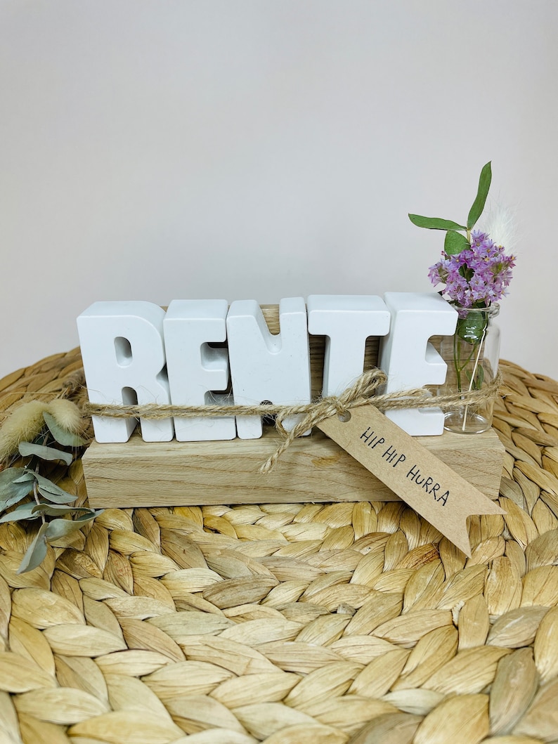 May include: White wooden letters spelling "RENTE" on a wooden base with a small vase of purple flowers and a tag that says "HIP HIP HURRA".