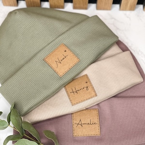 Könnte beinhalten: Drei gestrickte Mützen in verschiedenen Farben, übereinander gestapelt. Die Mützen haben einen braunen Leder-Patch mit einem Namen auf jedem. Die Namen sind Noah, Henry und Amelie.