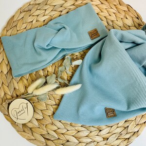 May include: A light blue ribbed knit headband and matching scarf set. Both items have a small brown leather tag with the text "Joneli handmade". The set is displayed on a woven straw mat with dried flowers.