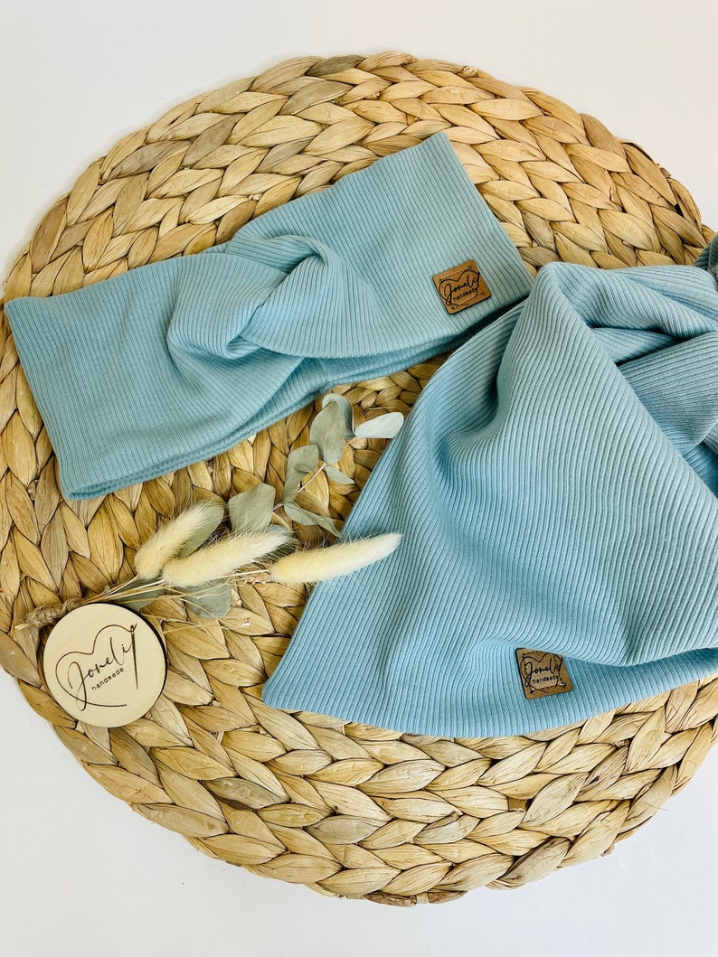 May include: A light blue ribbed knit headband and matching scarf set. Both items have a small brown leather tag with the text "Joneli handmade". The set is displayed on a woven straw mat with dried flowers.