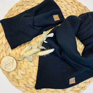 May include: Black ribbed knit headband and scarf set. The headband is twisted at the front. Both pieces have a small, square, brown leather tag with the text "Lovely Handmade". The set is displayed on a woven, circular mat.