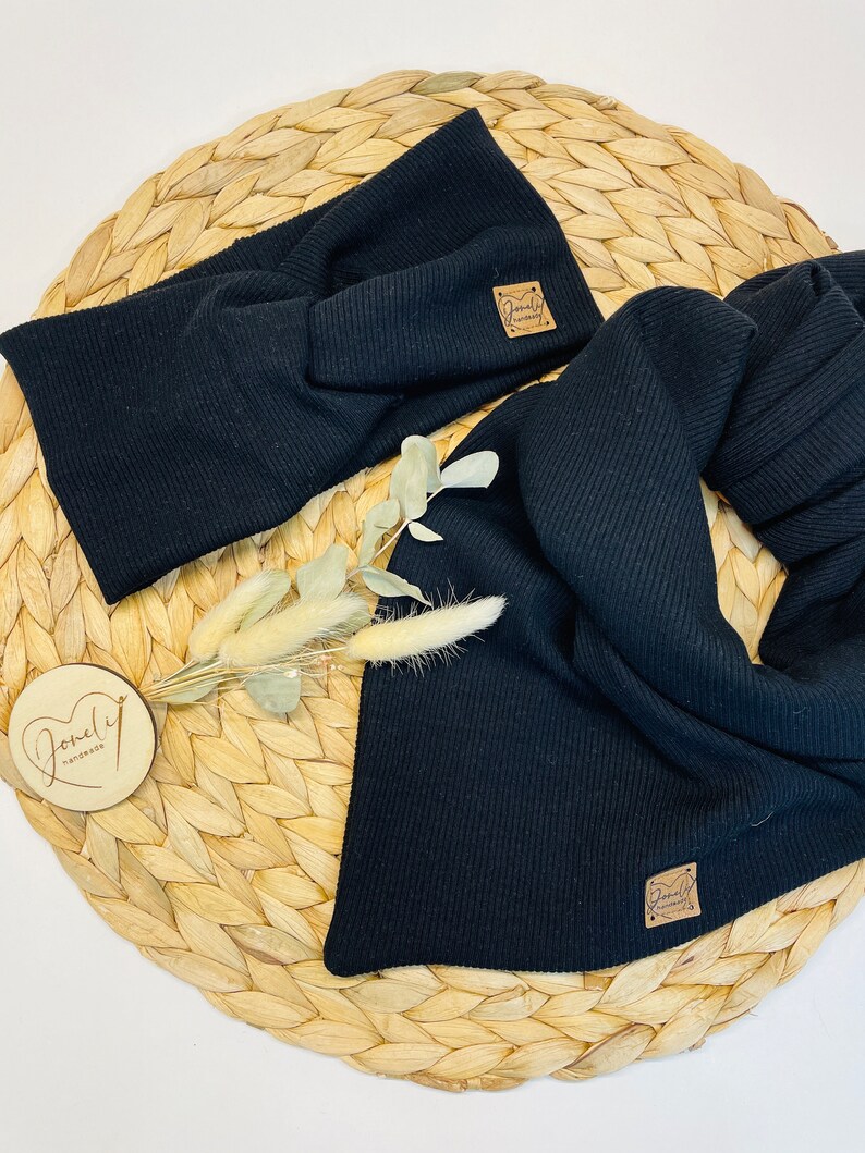 May include: Black ribbed knit headband and scarf set. The headband is twisted at the front. Both pieces have a small, square, brown leather tag with the text "Lovely Handmade". The set is displayed on a woven, circular mat.