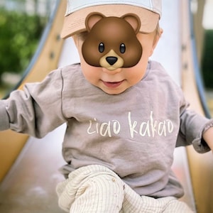 May include: A toddler wearing a light brown long-sleeved shirt with the text "ciao kakao" printed on the front. The toddler is also wearing a light brown and blue baseball cap with the text "Have a Great Day!" on the front. The toddler is sitting on a slide.