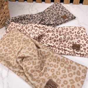 May include: Three leopard print headbands in tan, beige, and taupe. Each headband features a ribbed texture and a small, brown leather tag with the brand name. The headbands are arranged on a white marble surface.