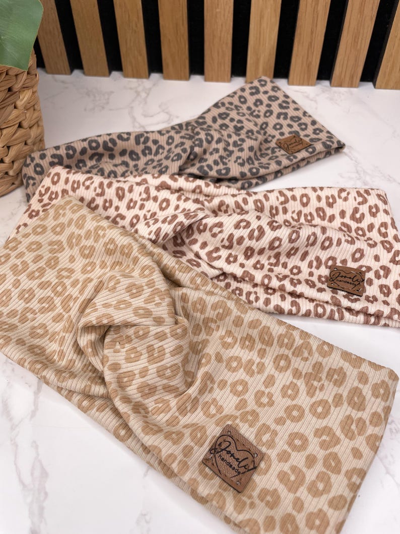 May include: Three leopard print headbands in tan, beige, and taupe. Each headband features a ribbed texture and a small, brown leather tag with the brand name. The headbands are arranged on a white marble surface.